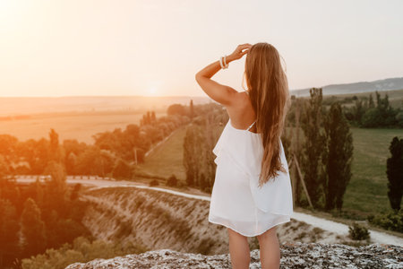 Happy woman standing with her back on the sunset in nature in summer with open hands. Romantic beautiful bride in white boho dress posing with mountains on sunsetの写真素材