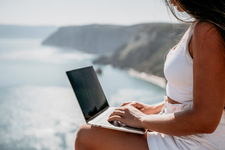 Digital nomad, woman in the hat, a business woman with a laptop sits on the rocks by the sea during sunset, makes a business transaction online from a distance. Freelance, remote work on vacation.の写真素材