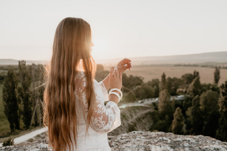 Happy woman in white boho dress on sunset in mountains. Romantic woman with long hair standing with her back on the sunset in nature in summer with open hands. Silhouette. Nature. Sunset.の写真素材
