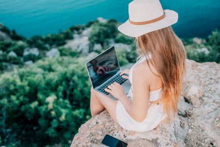 Woman laptop sea. Working remotely on seashore. Happy successful woman female freelancer in straw hat working on laptop by the sea at sunset. Freelance, remote work on vacationの写真素材