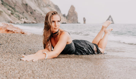 Woman summer travel sea. Happy tourist in black dress enjoy taking picture outdoors for memories. Woman traveler posing on sea beach surrounded by volcanic mountains, sharing travel adventure journeyの写真素材