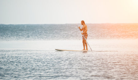 Sea man sup. Asian athlete man swimming in sea and paddleboarding at summer sunset. Healthy strong male enjoy outdoor active lifestyle and water sports on holiday vacation.の写真素材