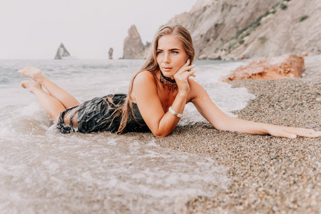 Woman summer travel sea. Happy tourist in black dress enjoy taking picture outdoors for memories. Woman traveler posing on sea beach surrounded by volcanic mountains, sharing travel adventure journeyの写真素材