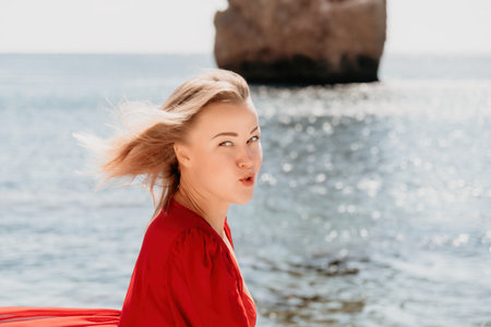 Woman summer travel sea. Happy tourist in long red dress enjoy taking picture outdoors for memories. Woman traveler posing on beach at sea surrounded by volcanic mountains, sharing travel adventureの写真素材