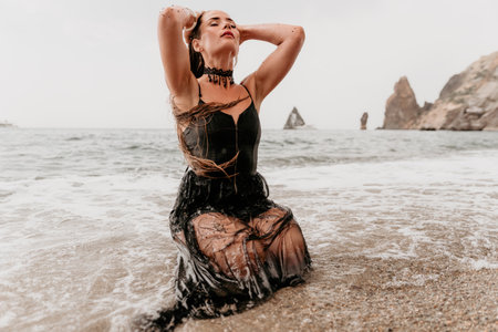 Woman summer travel sea. Happy tourist in black dress enjoy taking picture outdoors for memories. Woman traveler posing on sea beach surrounded by volcanic mountains, sharing travel adventure journeyの写真素材