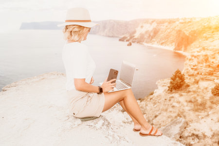 Digital nomad, Business woman working on laptop by the sea. Pretty lady typing on computer by the sea at sunset, makes a business transaction online from a distance. Freelance, remote work on vacationの写真素材
