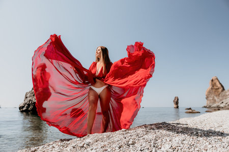Woman travel sea. Happy tourist in red dress enjoy taking picture outdoors for memories. Woman traveler posing on the rock at sea bay surrounded by volcanic mountains, sharing travel adventure journeyの写真素材