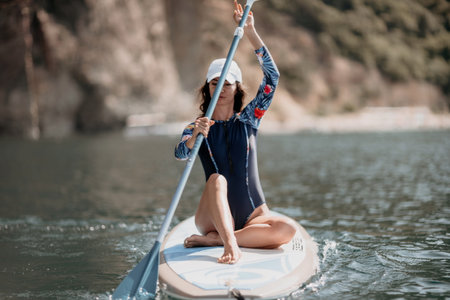Woman sup yoga. Middle age sporty woman practising yoga pilates on paddle sup surfboard. Female stretching doing workout on sea water. Modern individual female hipster outdoor summer sport activity.の写真素材