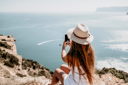Digital nomad, woman in the hat, a business woman with a laptop sits on the rocks by the sea during sunset, makes a business transaction online from a distance. Freelance, remote work on vacation.の写真素材