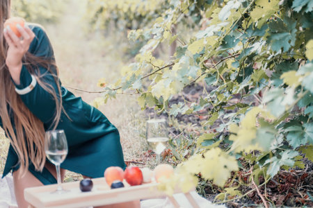 Woman picnic vineyard. Happy woman with a glass of wine at a picnic in the vineyard, wine tasting at sunset and open nature in the summer. Romantic dinner, fruit and wine.の写真素材
