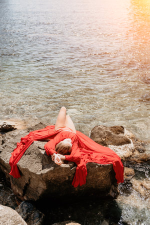 Woman summer travel sea. Happy tourist in long red dress enjoy taking picture outdoors for memories. Woman traveler posing on beach at sea surrounded by volcanic mountains, sharing travel adventureの写真素材