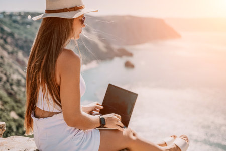 Digital nomad, woman in the hat, a business woman with a laptop sits on the rocks by the sea during sunset, makes a business transaction online from a distance. Freelance, remote work on vacation.の写真素材