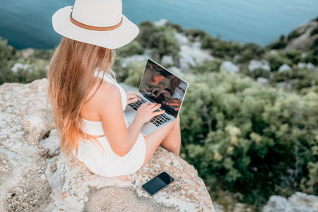 Woman laptop sea. Working remotely on seashore. Happy successful woman female freelancer in straw hat working on laptop by the sea at sunset. Freelance, remote work on vacationの写真素材