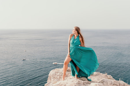 Woman travel portrait. Happy woman with long hair looking at camera and smiling. Close up portrait cute woman in a mint long dress posing on a volcanic rock high above the seaの写真素材
