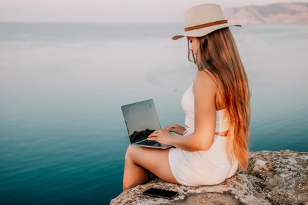 Woman laptop sea. Working remotely on seashore. Happy successful woman female freelancer in straw hat working on laptop by the sea at sunset. Freelance, remote work on vacationの写真素材