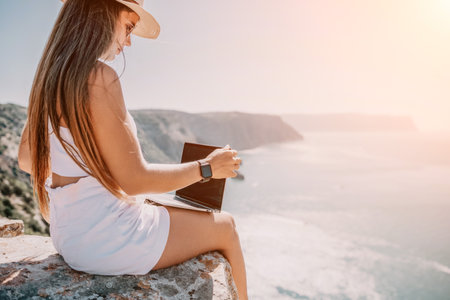 Digital nomad, woman in the hat, a business woman with a laptop sits on the rocks by the sea during sunset, makes a business transaction online from a distance. Freelance, remote work on vacation.の写真素材