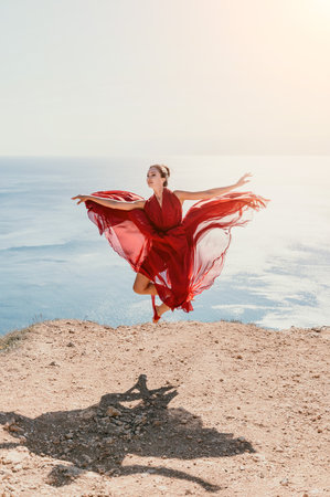 Woman in red dress on sea. Side view a Young beautiful sensual woman in a red long dress posing on a rock high above the sea on sunset. Girl on the nature on blue sky background. Fashion photo.の写真素材
