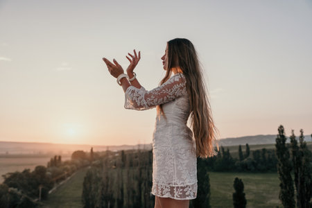 Happy woman in white boho dress on sunset in mountains. Romantic woman with long hair standing with her back on the sunset in nature in summer with open hands. Silhouette. Nature. Sunset.の写真素材