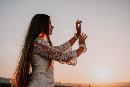 Happy woman in white boho dress on sunset in mountains. Romantic woman with long hair standing with her back on the sunset in nature in summer with open hands. Silhouette. Nature. Sunset.の写真素材