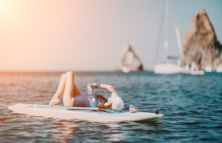 Woman sup yoga. Middle age sporty woman practising yoga pilates on paddle sup surfboard. Female stretching doing workout on sea water. Modern individual female hipster outdoor summer sport activity.の写真素材