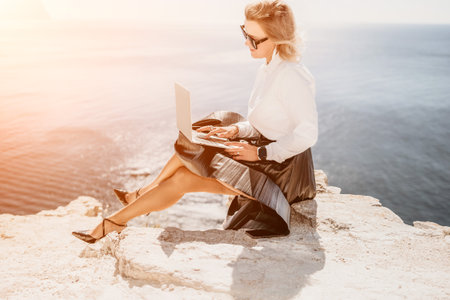 Digital nomad, Business woman working on laptop by the sea. Pretty lady typing on computer by the sea at sunset, makes a business transaction online from a distance. Freelance, remote work on vacationの写真素材