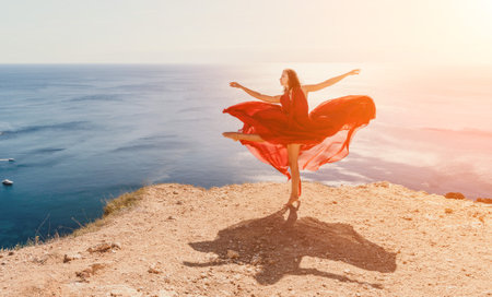 Woman in red dress on sea. Side view a Young beautiful sensual woman in a red long dress posing on a rock high above the sea on sunset. Girl on the nature on blue sky background. Fashion photo.の写真素材