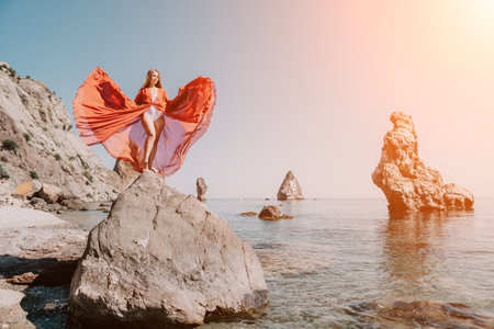 Woman travel sea. Young Happy woman in a long red dress posing on a beach near the sea on background of volcanic rocks, like in Iceland, sharing travel adventure journeyの写真素材