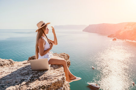 Woman laptop sea. Working remotely on seashore. Happy successful woman female freelancer in straw hat working on laptop by the sea at sunset. Freelance, remote work on vacationの写真素材