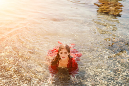 Woman travel sea. Happy tourist in red dress enjoy taking picture outdoors for memories. Woman traveler posing in sea beach, surrounded by volcanic mountains, sharing travel adventure journeyの写真素材