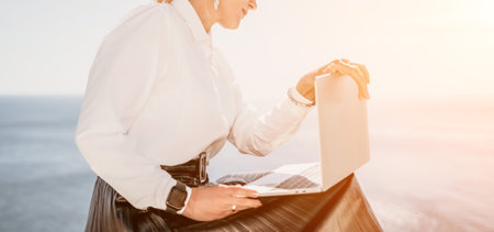 Digital nomad, Business woman working on laptop by the sea. Pretty lady typing on computer by the sea at sunset, makes a business transaction online from a distance. Freelance, remote work on vacationの写真素材