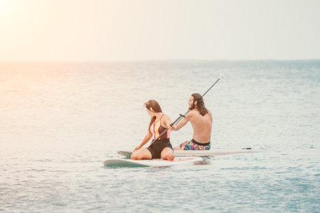 Sea woman and man on sup. Silhouette of happy young woman and man, surfing on SUP board, confident paddling through water surface. Idyllic sunset. Active lifestyle at sea or river.の写真素材