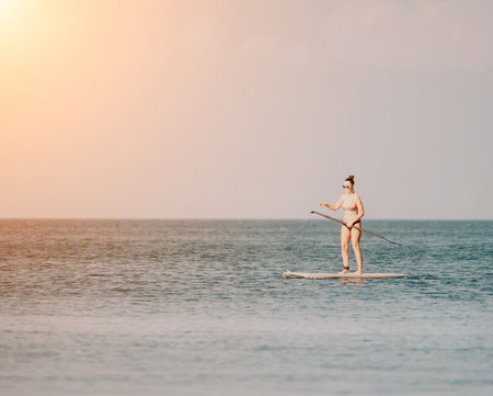 Sea woman sup. Silhouette of happy middle aged tanned woman in rainbow bikini, surfing on SUP board, confident paddling through water surface. Idyllic sunset. Active lifestyle at sea or river.の写真素材
