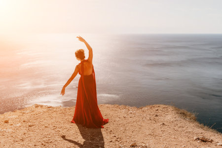 Woman in red dress on sea. Side view a Young beautiful sensual woman in a red long dress posing on a rock high above the sea on sunset. Girl on the nature on blue sky background. Fashion photo.の写真素材