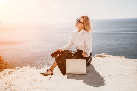Digital nomad, Business woman working on laptop by the sea. Pretty lady typing on computer by the sea at sunset, makes a business transaction online from a distance. Freelance, remote work on vacationの写真素材