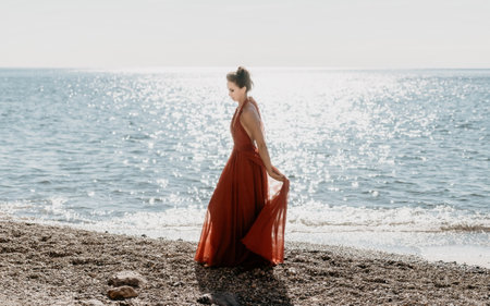 Woman in red dress on sea. Side view a Young beautiful sensual woman in a red long dress posing on the beach near sea on sunset. Girl on the nature on blue sky background. Fashion photo.の写真素材