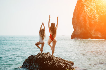 Woman and her daughter practicing balancing yoga pose on one leg up together on rock in the sea. Silhouette mother and daughter doing yoga at beachの写真素材