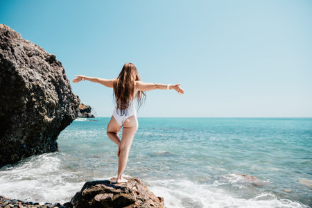 Woman sea yoga. Back view of free calm happy satisfied woman with long hair standing on top rock with yoga position against of sky by the sea. Healthy lifestyle outdoors in nature, fitness concept.の写真素材