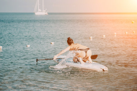 Sea woman sup. Silhouette of happy middle aged tanned woman in rainbow bikini, surfing on SUP board, confident paddling through water surface. Idyllic sunset. Active lifestyle at sea or river.の写真素材