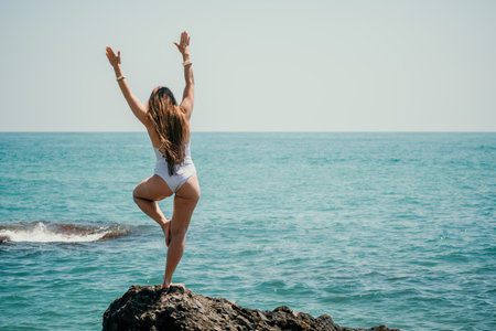 Woman sea yoga. Back view of free calm happy satisfied woman with long hair standing on top rock with yoga position against of sky by the sea. Healthy lifestyle outdoors in nature, fitness concept.の写真素材