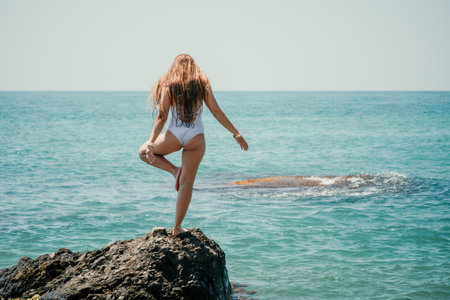 Woman sea yoga. Back view of free calm happy satisfied woman with long hair standing on top rock with yoga position against of sky by the sea. Healthy lifestyle outdoors in nature, fitness concept.の写真素材