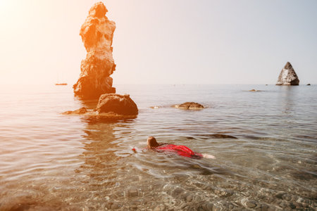 Woman travel sea. Happy tourist in red dress enjoy taking picture outdoors for memories. Woman traveler posing in sea beach, surrounded by volcanic mountains, sharing travel adventure journeyの写真素材