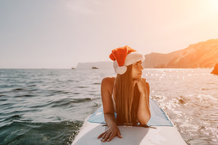Woman sea sup. Close up portrait of happy young caucasian woman with long hair in Santa hat looking at camera and smiling. Cute woman portrait in a white bikini posing on sup board in the seaの写真素材