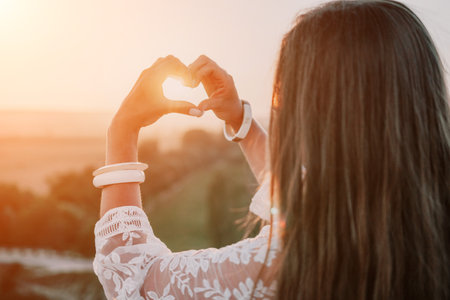 Happy woman in white boho dress making heart sign with hands on sunset in mountains. Romantic woman with long hair standing with her back on the sunset in nature in summer with open hands.の写真素材