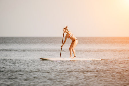 Sea woman sup. Happy woman in a rainbow-coloured bikini stands on SUP board. She is paddling through the calm water, creating ripples along the way. Idyllic sunset. Active lifestyle at sea or river.の写真素材