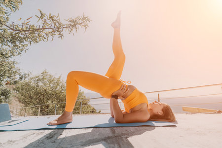 Fitness woman sea. Happy middle aged woman in orange sportswear exercises morning outdoors on yoga mat with laptop in park over ocean beach. Female fitness pilates yoga routine. Healthy lifestyle.の写真素材