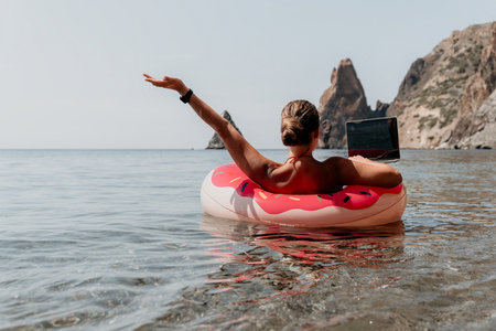 Woman freelancer works on laptop swimming in sea on pink inflatable ring. Happy tourist floating on...