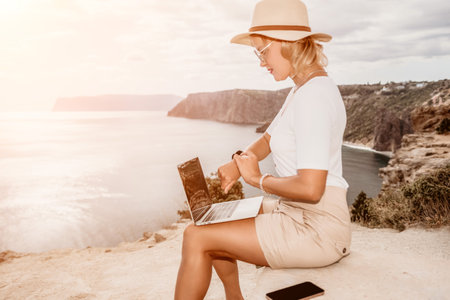 Digital nomad, Business woman working on laptop by the sea. Pretty lady typing on computer by the sea at sunset, makes a business transaction online from a distance. Freelance, remote work on vacationの写真素材
