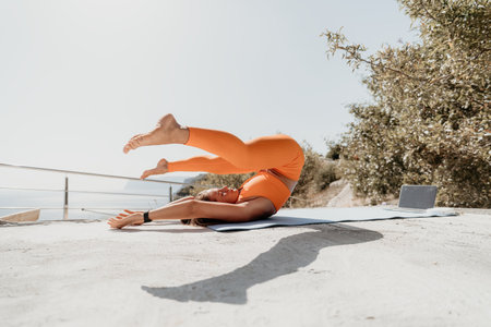 Fitness woman sea. Happy middle aged woman in orange sportswear exercises morning outdoors on yoga mat with laptop in park over ocean beach. Female fitness pilates yoga routine. Healthy lifestyle.の写真素材