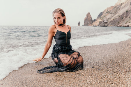 Woman summer travel sea. Happy tourist in black dress enjoy taking picture outdoors for memories. Woman traveler posing on sea beach surrounded by volcanic mountains, sharing travel adventure journeyの写真素材