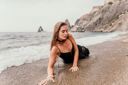Woman summer travel sea. Happy tourist in black dress enjoy taking picture outdoors for memories. Woman traveler posing on sea beach surrounded by volcanic mountains, sharing travel adventure journeyの写真素材